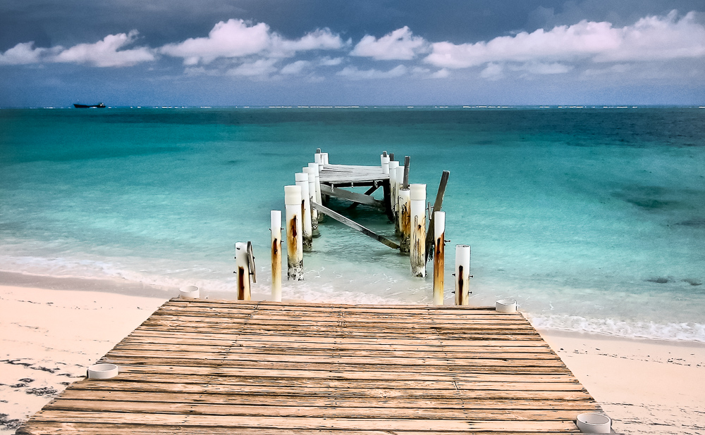 Color photograph: A small derelict wooden pier juts straight out into ocean. The background is void of all elements except the natural seascape. It transitions from white sand, azure waters, deep blue ocean, a single low band of distinctly formed clouds, and finally to a deep blue sky.